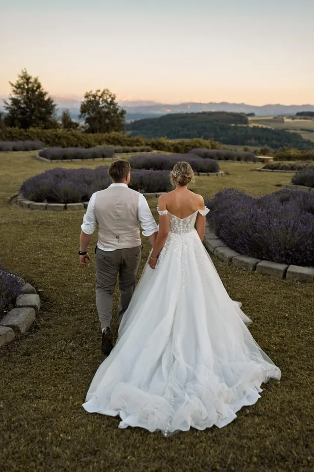 Brautpaar Hand in Hand auf einer Wiese mit Lavendel bei Sonnenuntergang, von hinten gesehen.