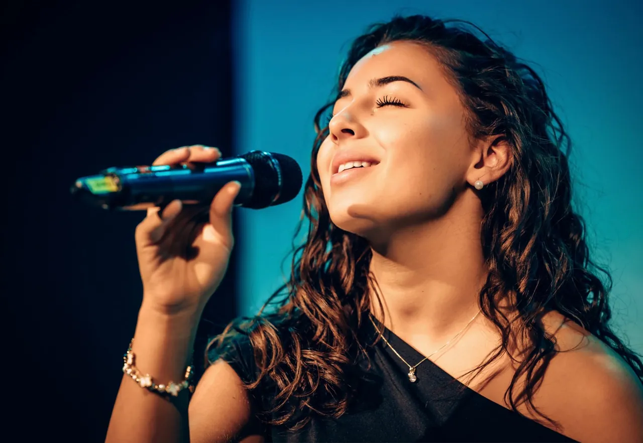 Shirin singt lächelnd mit geschlossenen Augen ins Mikrofon, vor türkisblauem Licht.